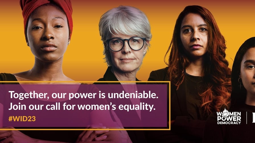 Four women standing in front of an orange background. The text in front of them says "Together, our power is undeniable. Join the call for women's equality. #WID23." 