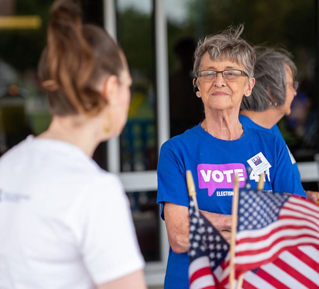 A woman in a VOTE411 T-Shirt speaking to another woman next to small American flags