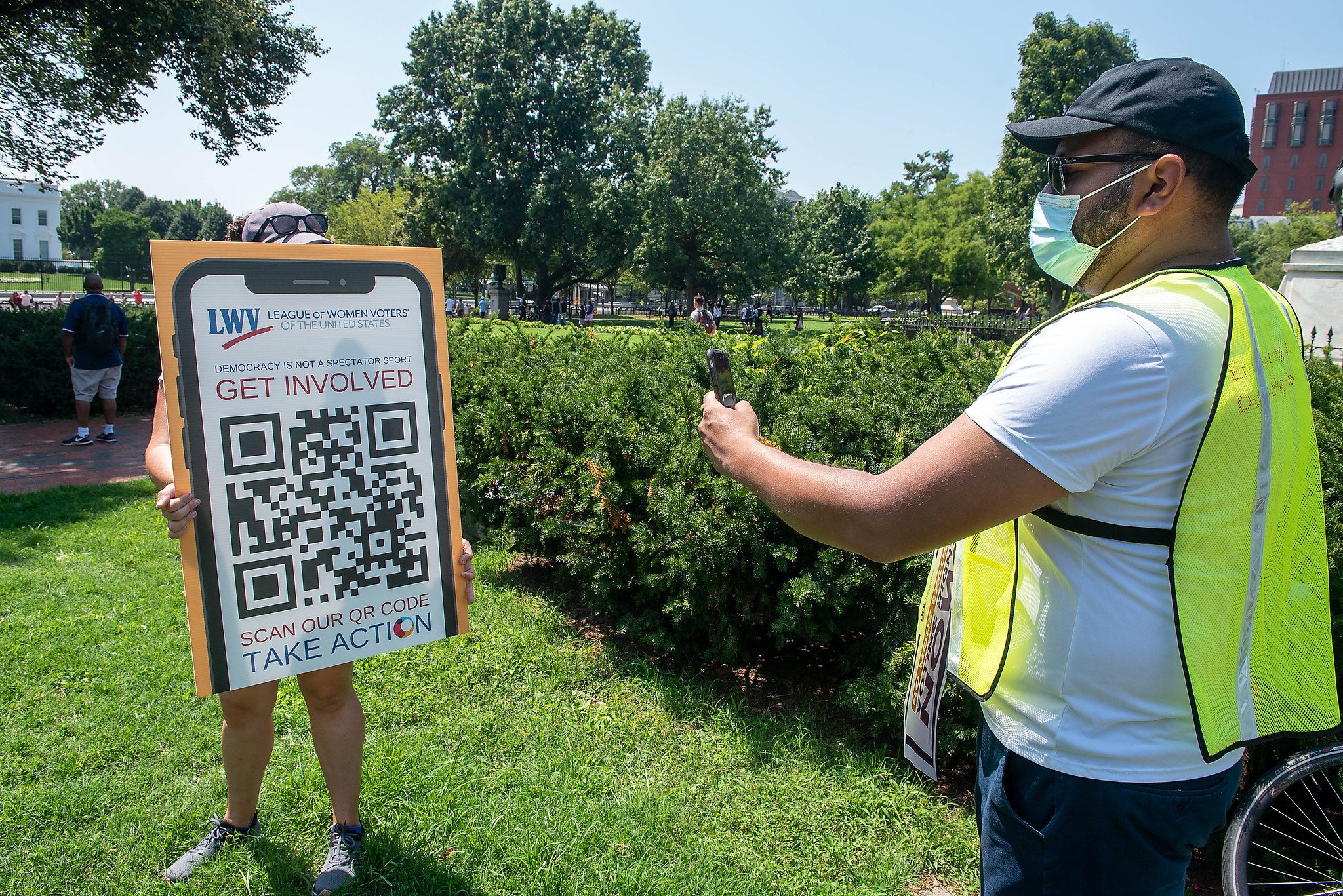 LWVUS staff holding up a poster board at a rally outside with a QR code to download OutreachCircle. On the right, a member of the rally is scanning the QR code on their smartphone.