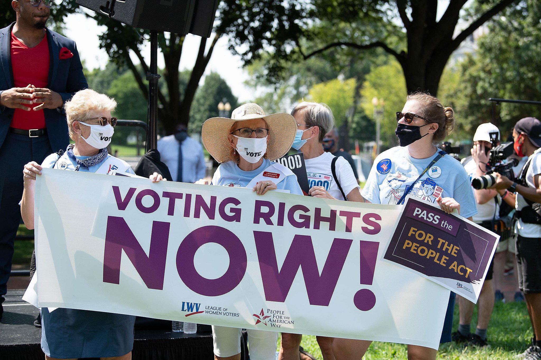 Three League members holding a white "VOTING RIGHTS NOW" banner at a rally in Washington, DC. The League members are wearing sunglasses and masks.