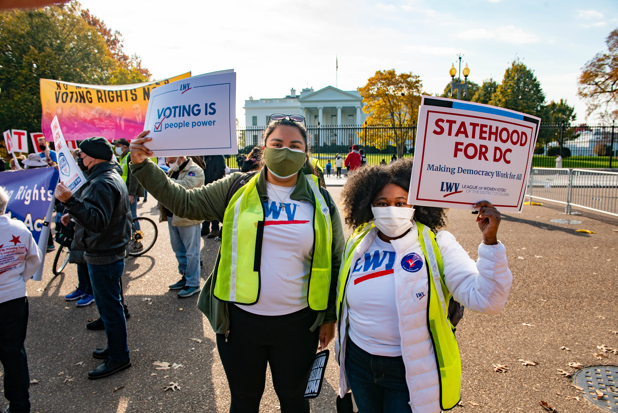 Two LWVUS staff members at a rally in front of the White House in Washington DC. Both staff members are wearing yellow vest and holding rally signs ("Voting is People Power," "STATEHOOD for DC")