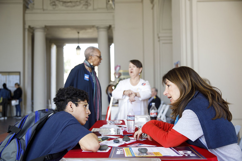 Man kneeling in front of a table covered in pamphlets, talking to a woman sitting behind the table