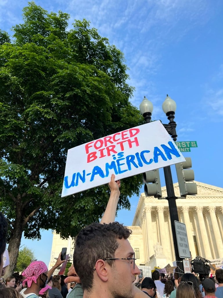 A person holding a sign in front of the Supreme Court that says "Forced Birth is Un-American"