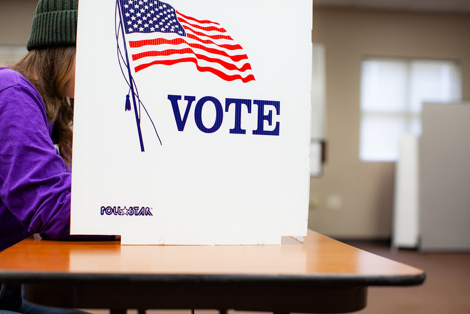 A person is voting behind a white sign with an American flag that says 