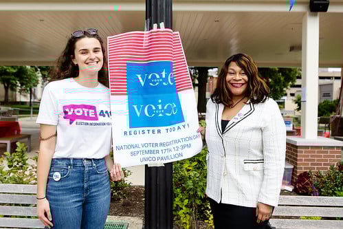 Two women, one wearing a VOTE411 shirt, standing next to a sign promoting voter registration
