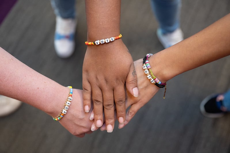 Three hands joined together, each with a "VOTE" bracelet on their wrists