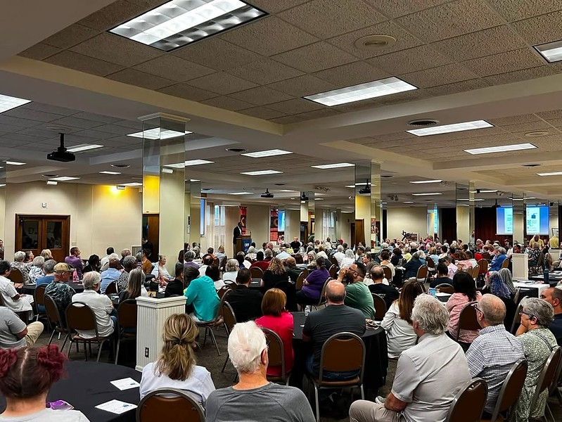 People sit in a crowded room listening to speakers