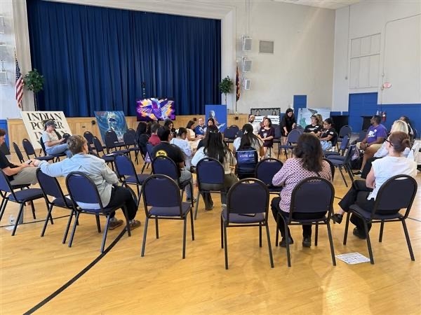 A crowd of people sitting in a circle with six works of art on stands behind them