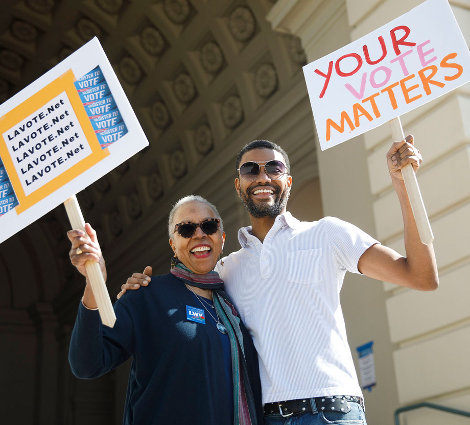 An older woman holding a sign, side hugging a young man also holding a sign that reads