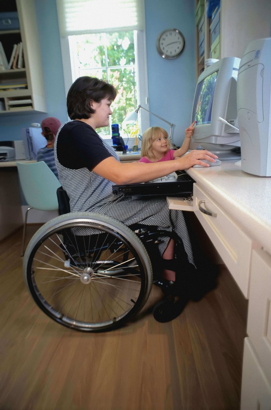 A woman in a wheelchair typing on a computer while a child watches