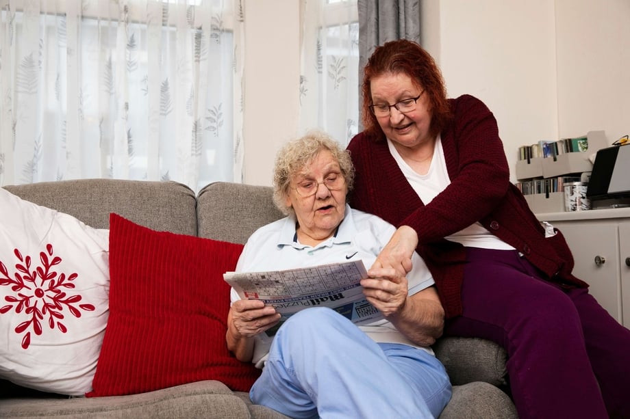 Two older women sitting on a couch looking at a newspaper
