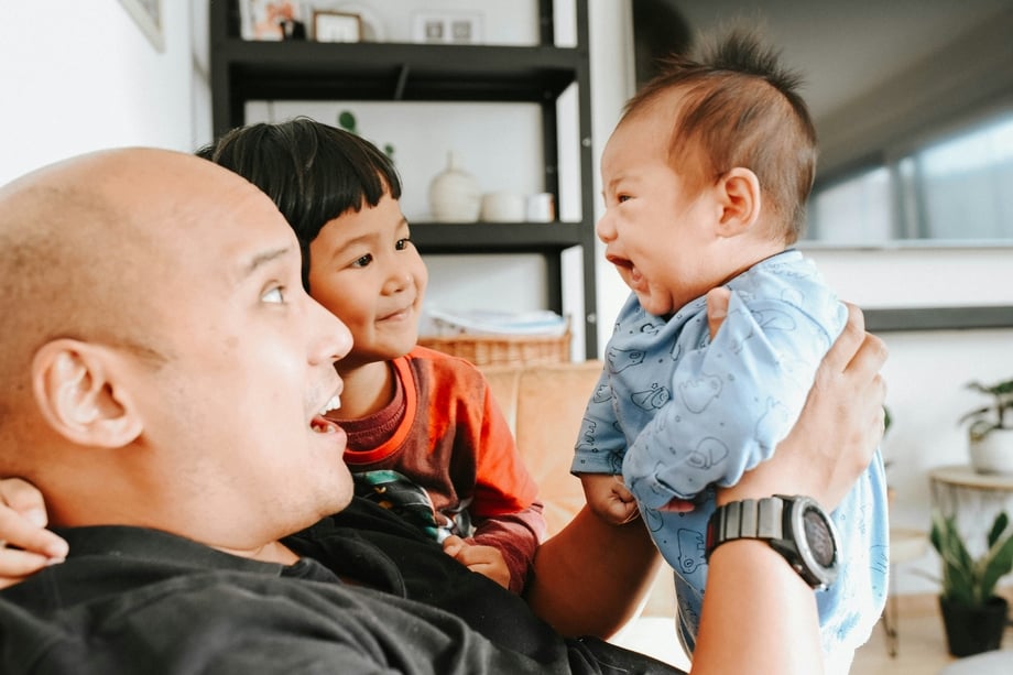 A caregiver sitting next to one child while holding up a smiling baby