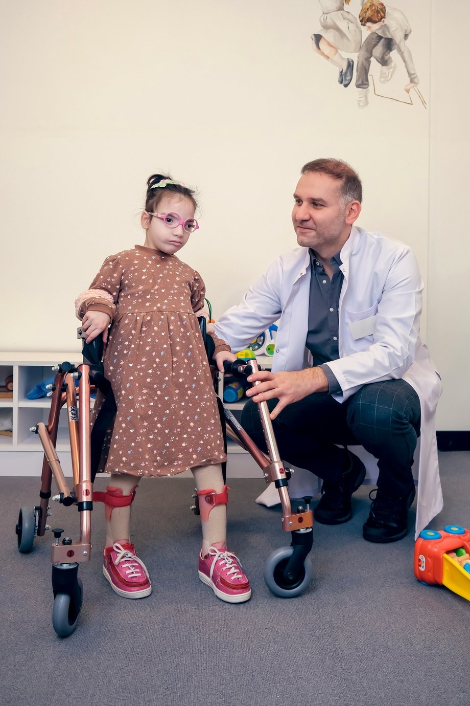 A girl using an assistive walking device standing next to her doctor, who is kneeling