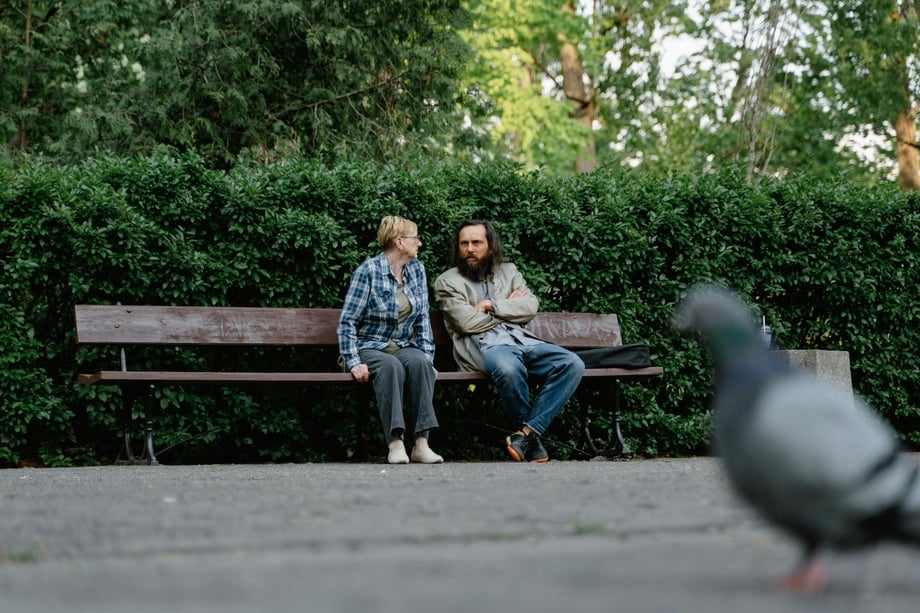 A woman and man sitting on a bench with a pigeon in the foreground