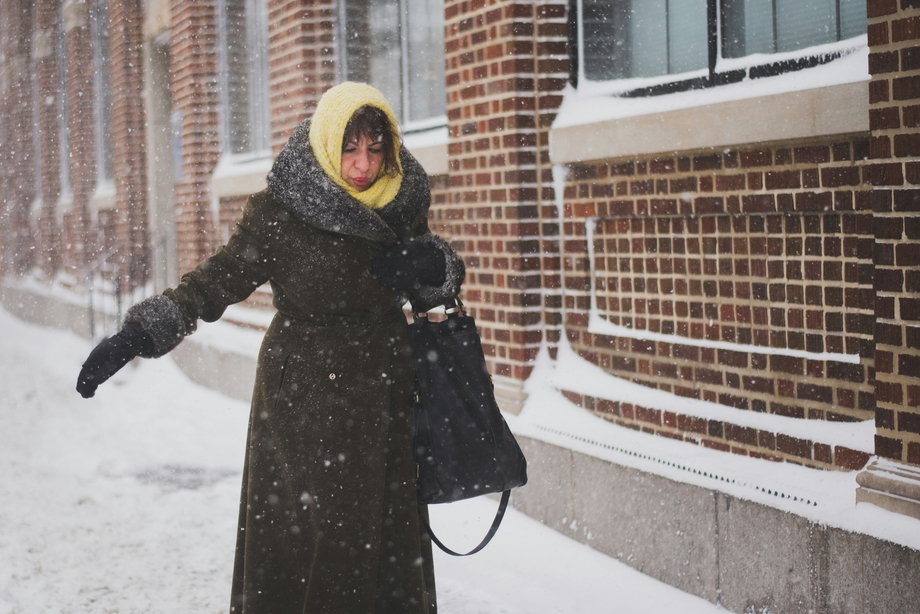 A woman in a black coat walking in the snow by a brick building